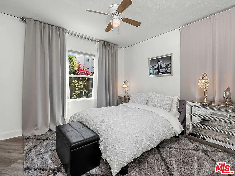 A bright bedroom at 8823 Betty Way with a ceiling fan, large window framing greenery, and warm wood floors